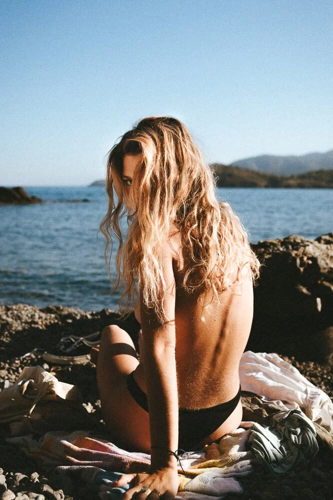 A woman with long, wavy blonde hair sitting topless on a towel by a rocky beach, facing away from the camera, with a view of water and mountains in the background during daytime.