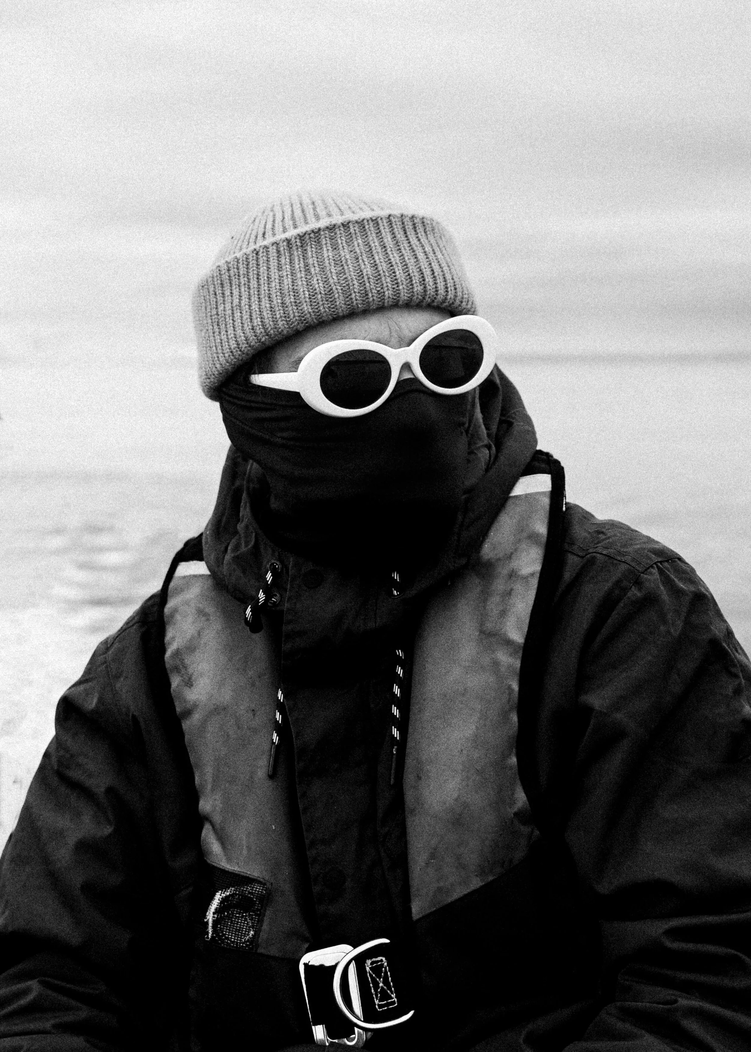 A person wearing a knitted beanie, large round sunglasses, a face covering, a jacket, and a backpack, holding a glass of beer, outdoors in a snowy environment.