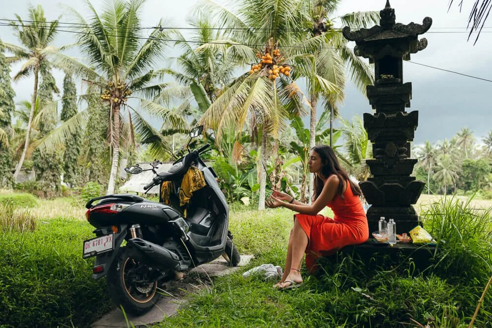 A woman in an orange dress sitting on a stone platform beside a black scooter and a black shrine, surrounded by tropical palm trees.