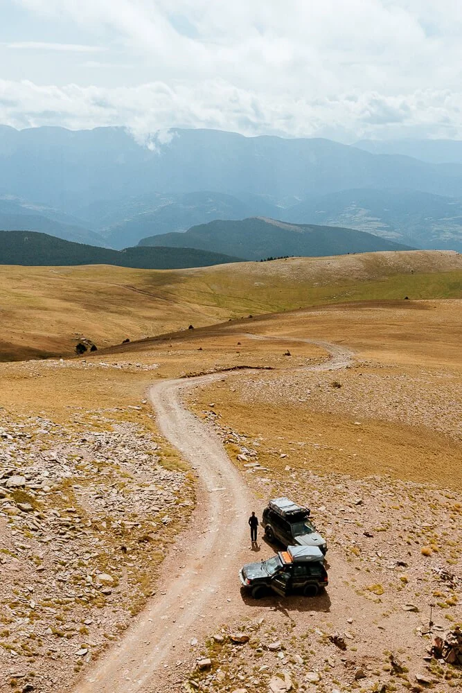 A person standing beside two off-road vehicles on a dirt trail through an open, rocky landscape with distant mountains and cloudy sky.
