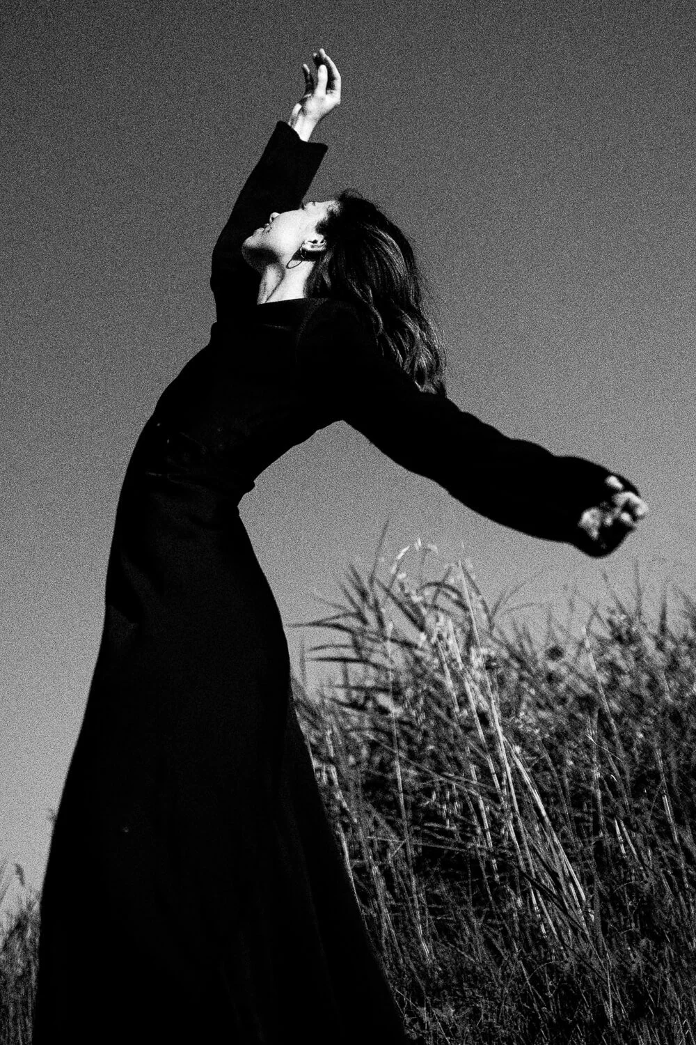 A woman in a black dress standing in a field of tall grass, arms raised, her head tilted back, captured in black and white.