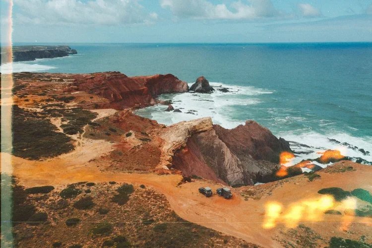 Côtes rocheuses de couleur rouge et orange avec mer agitée et deux véhicules garés sur un chemin de terre.