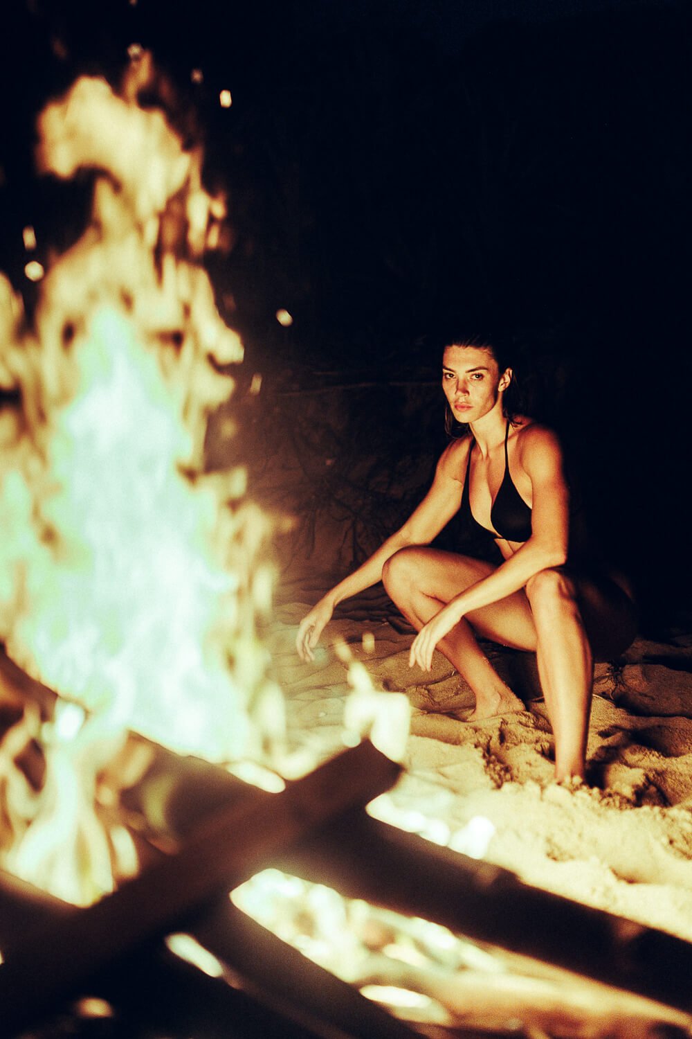A woman in a black bikini crouching on sandy ground near a campfire at night.