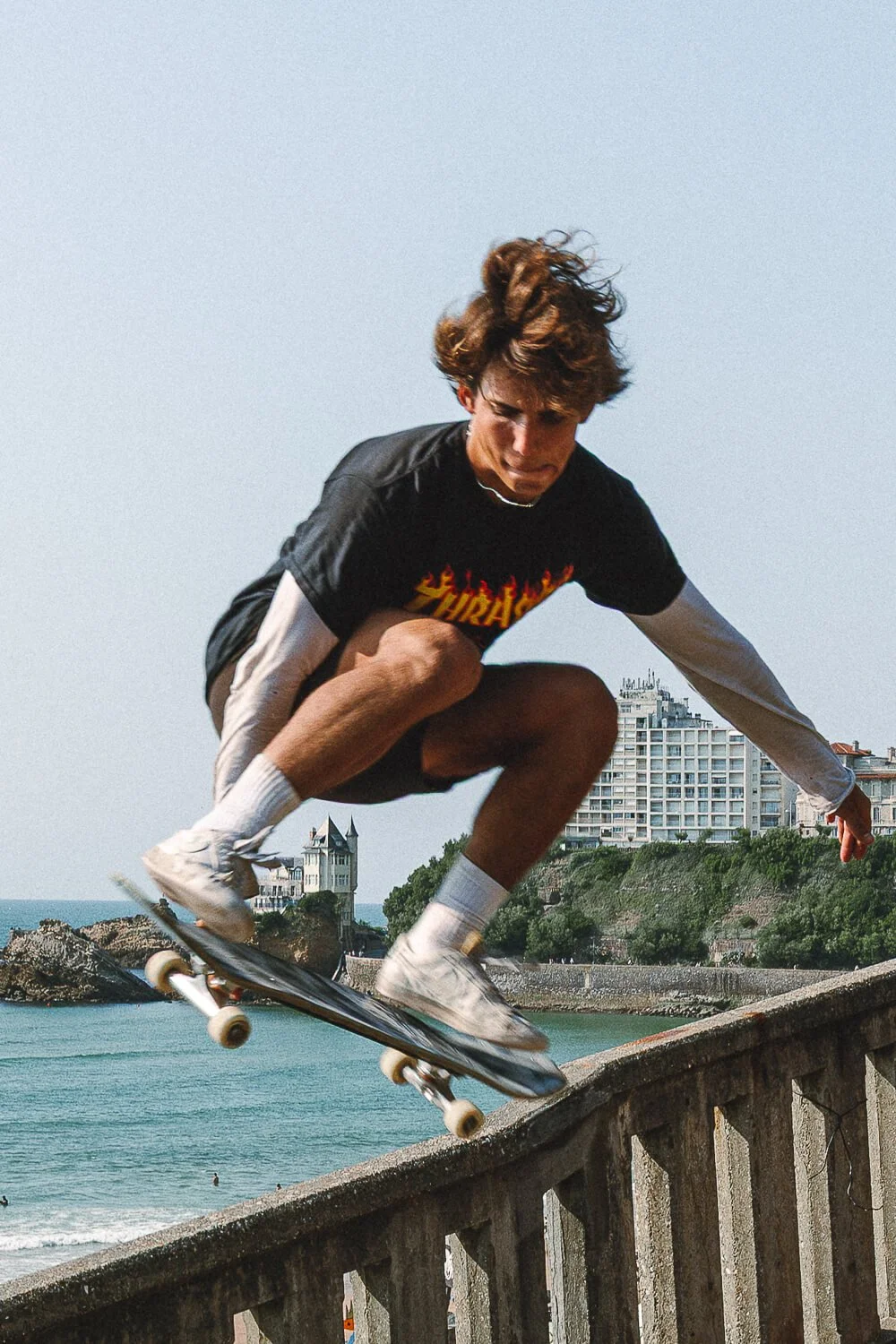 A young man skateboarding on a concrete ledge near the beach, with buildings and the ocean in the background on a clear day.
