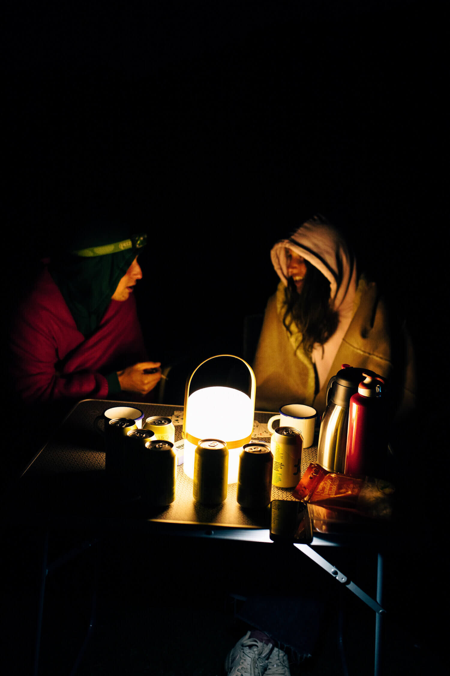 Deux personnes assises à une table de camping, éclairées par une lanterne. Des canettes, des tasses et des thermos sont posés sur la table.