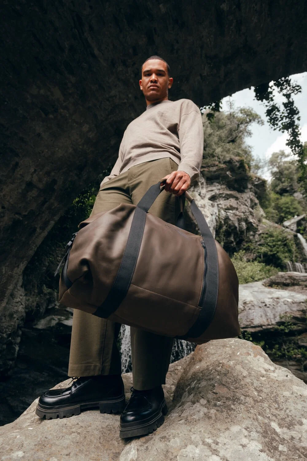 A man standing on a large rock outdoors, holding a brown duffle bag, with a natural rocky landscape, trees, and waterfall in the background.