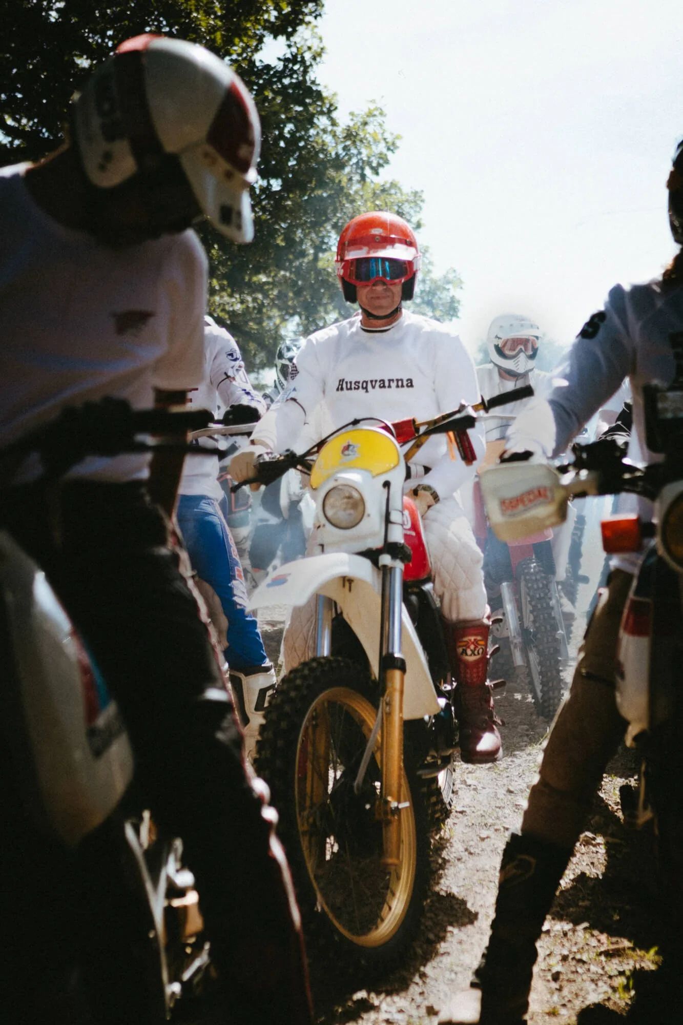 Group of motocross riders wearing helmets and protective gear, gathered around dirt bikes outdoors in a wooded area.