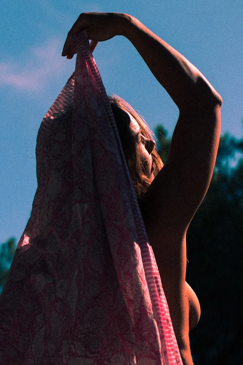 A woman with long hair standing in sunlight with her eyes closed, holding a patterned cloth above her head, against a background of a clear blue sky and trees.