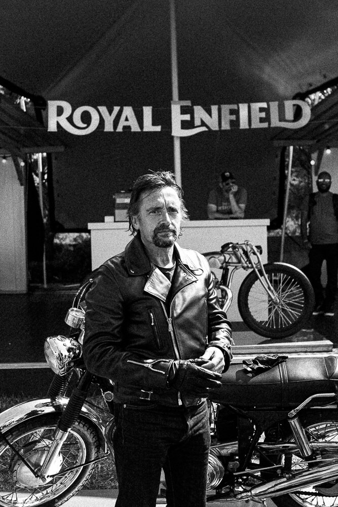 A man in a leather jacket standing in front of a vintage motorcycle at the Royal Enfield booth, with a bicycle displayed behind him and a large Royal Enfield sign overhead.