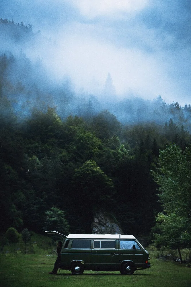 A person standing next to a green van with an open door in a grassy clearing, surrounded by lush green trees and a misty, fog-covered mountain landscape.