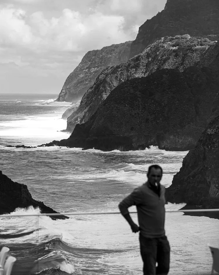 Un homme debout sur une plage rocailleuse, avec des falaises escarpées en arrière-plan, vue en noir et blanc.