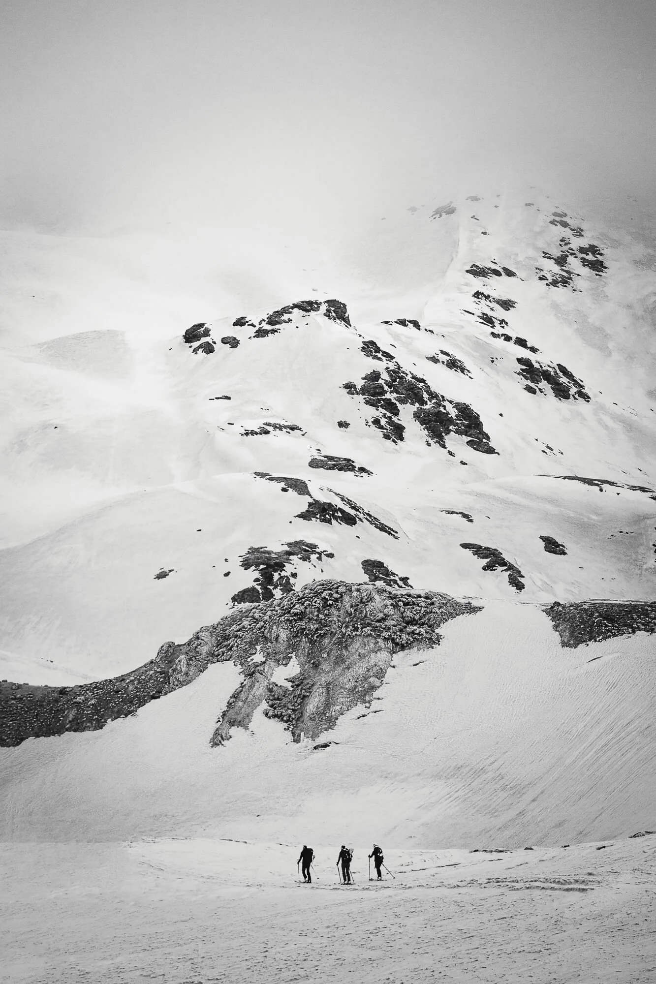 Three hikers dressed in winter gear walking in a snowy landscape under a steep, snow-covered mountain with dark rock patches and misty sky.