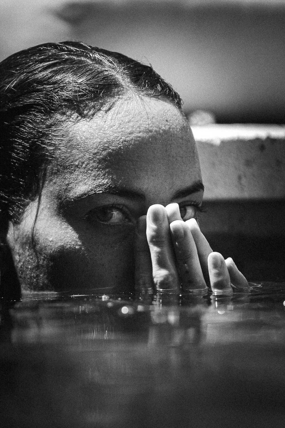 A close-up black and white photo of a person partially submerged in water, with their face partly visible above the surface and hand touching their face.