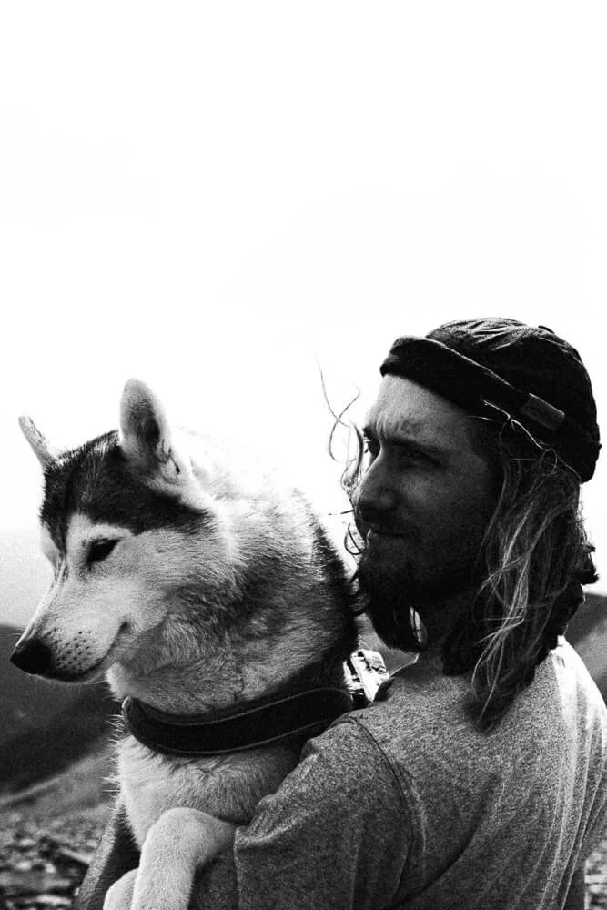 A man with long hair wearing a headband holds a husky dog outdoors in black and white.