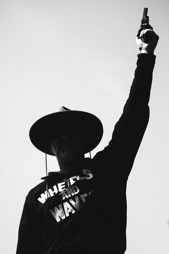 Silhouette of a person wearing a wide-brimmed hat, sunglasses, and a jacket with the text "WHEELS AND WAVES", holding a gun raised in the air against a plain background.