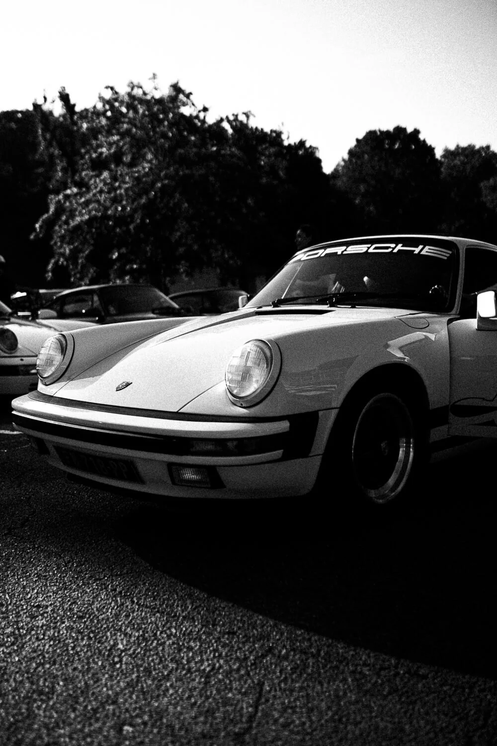 Black and white photo of a classic Porsche sports car parked outdoors with trees in the background.