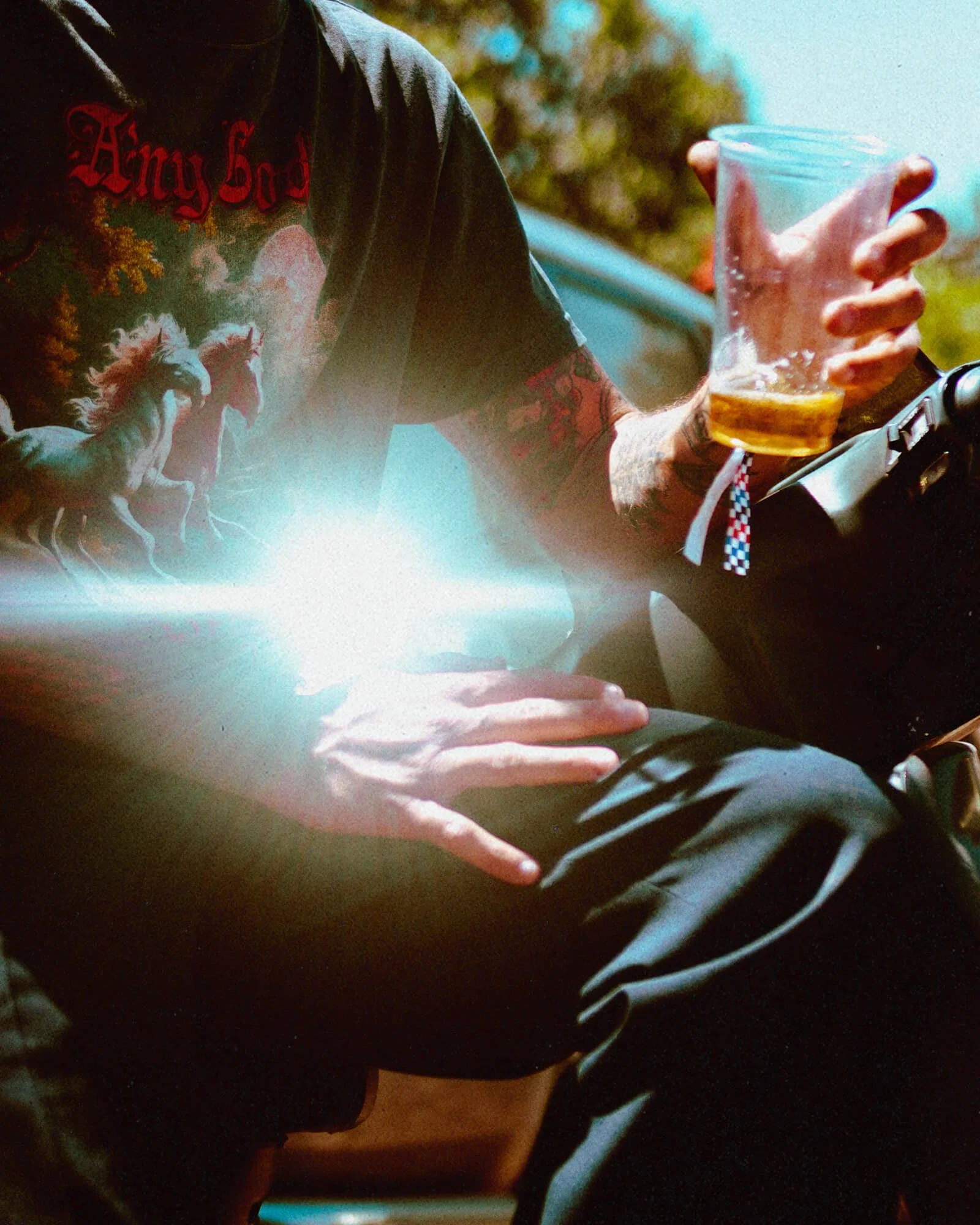 Person holding a clear glass mug with a beverage, wearing a black T-shirt with red writing and graphic, tattooed arm, and a wristband with a red, white, and blue pattern.