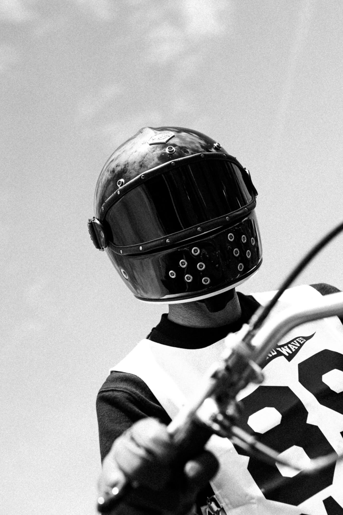 A person wearing a black helmet with a reflective visor, a white and black shirt, and riding a motorcycle.