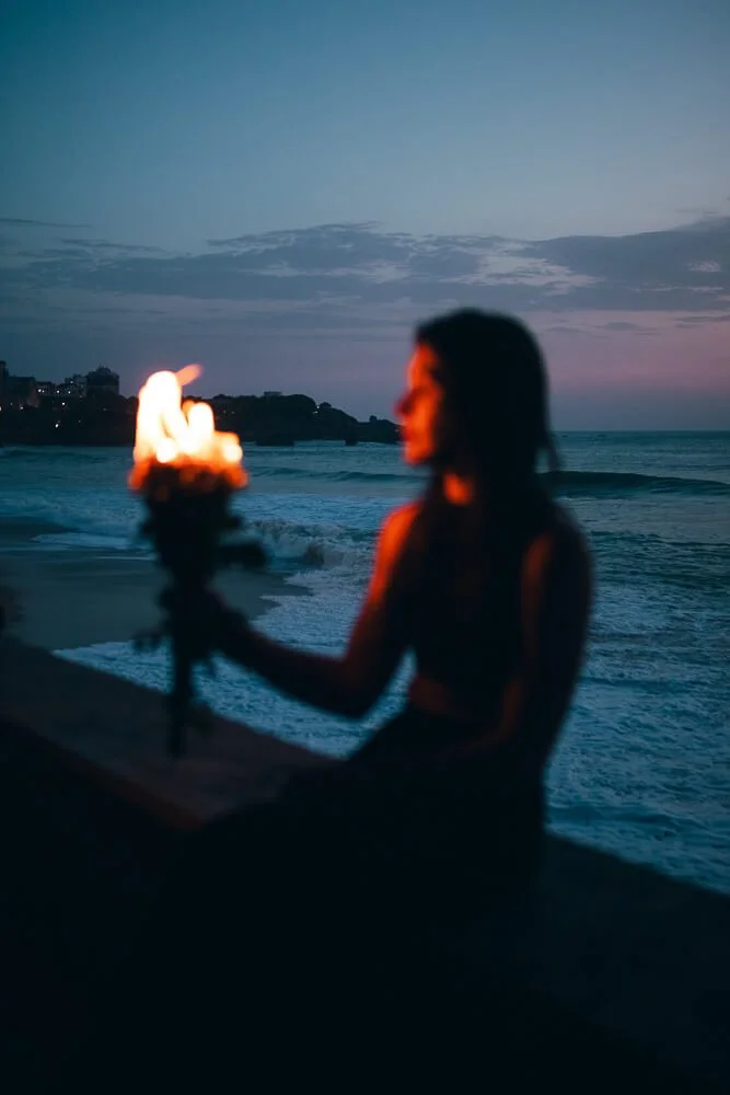 Silhouette of a woman at the beach holding a flaming torch during dusk, with waves and a cloudy sky in the background.