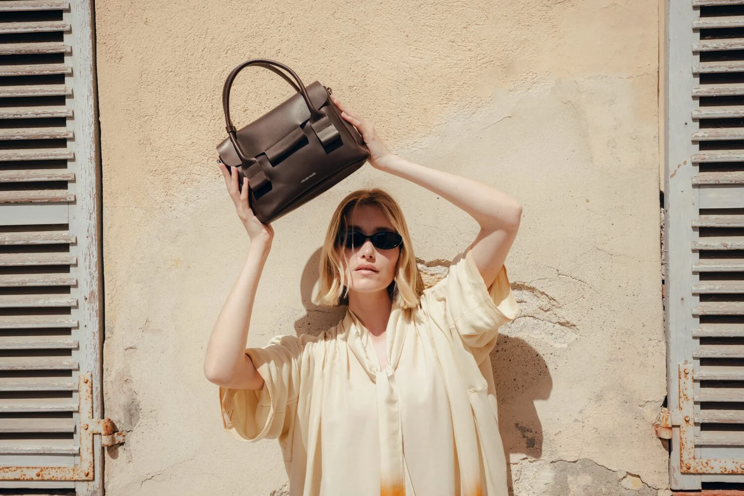 A woman with shoulder-length blonde hair, wearing dark sunglasses, a beige outfit, and holding a brown handbag above her head, standing against a textured beige wall with shutters on either side.