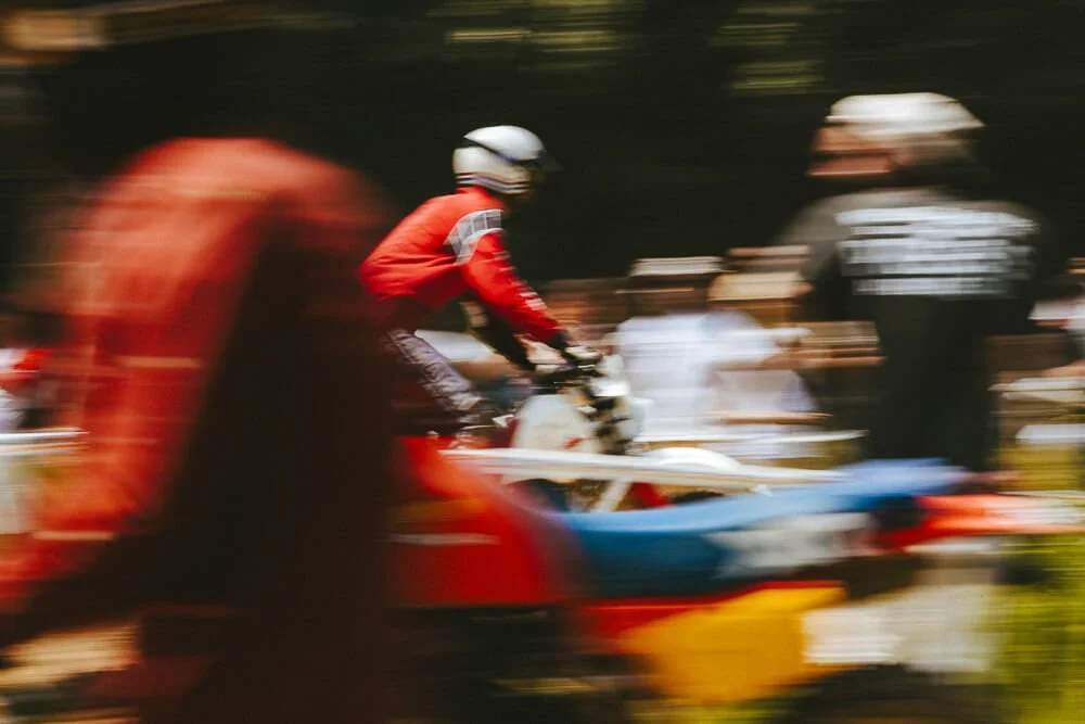 Blurred image of a person riding a jet ski or watercraft with a blue, white, and red body, surrounded by other people, some wearing helmets, outdoors during daytime.