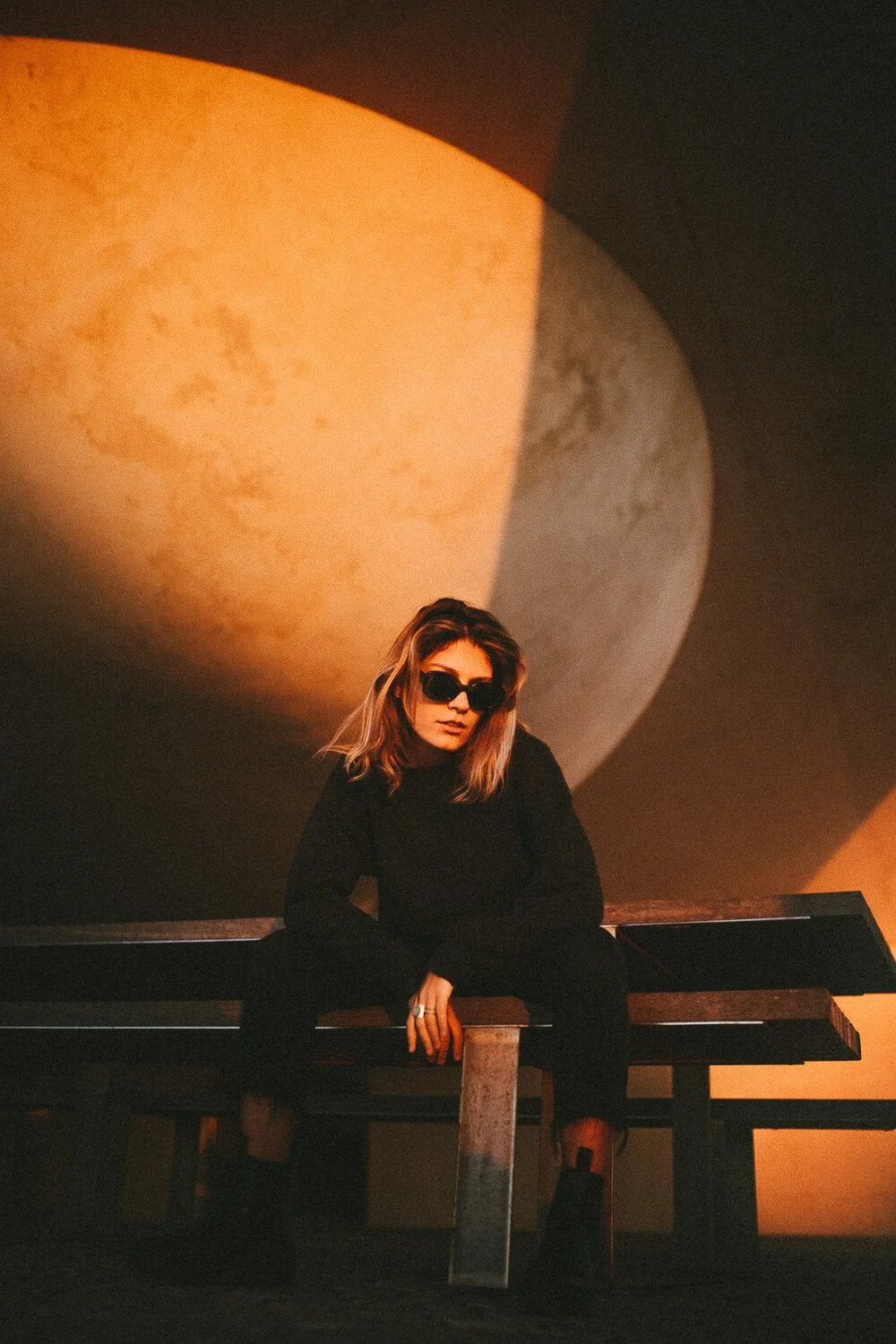 A woman with sunglasses sitting on a wooden bench in front of a large illuminated moon backdrop.