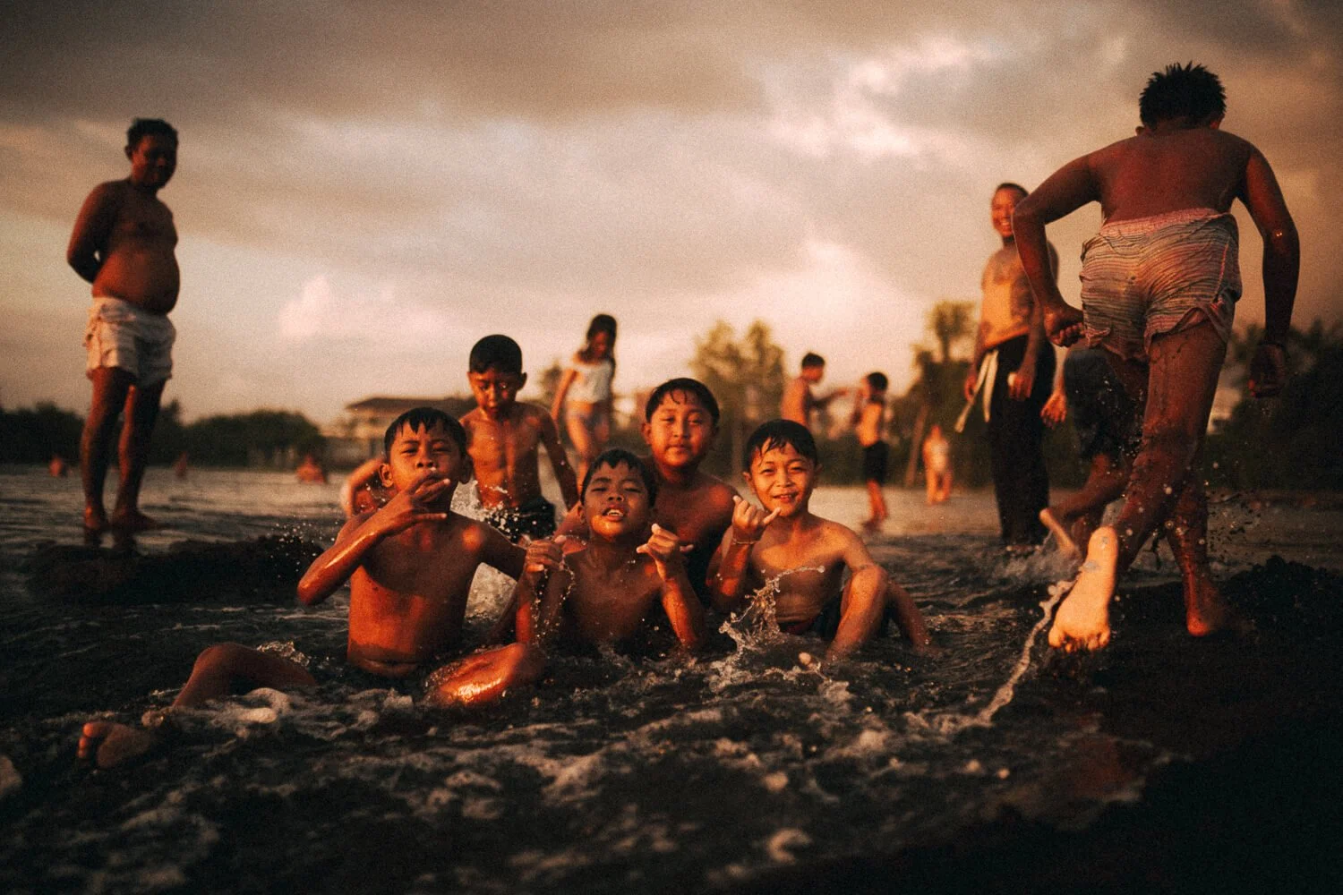 Des enfants jouent et nagent dans une rivière au coucher du soleil, avec un ciel coloré en arrière-plan.