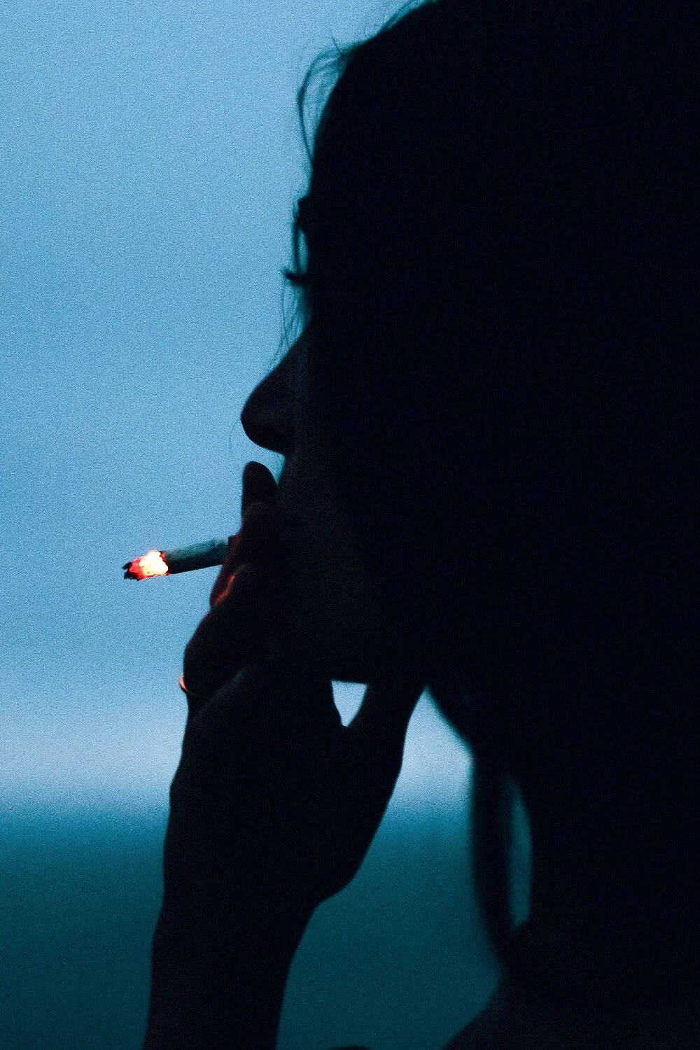 Silhouette of a person smoking a cigarette against a blue sky.