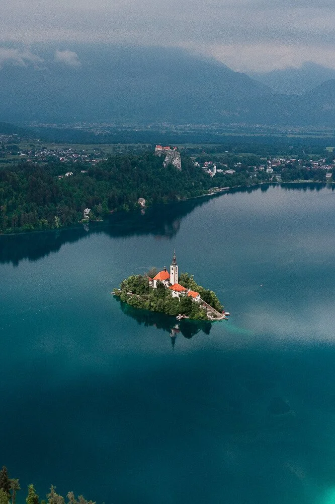 Aerial view of Bled Island with a church and red-roofed buildings on Lake Bled in Slovenia, with mountains in the background.