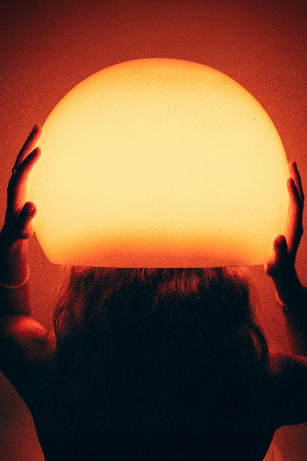 Person holding a large round glowing orange lamp that illuminates the person's head and hands, against a dark background.