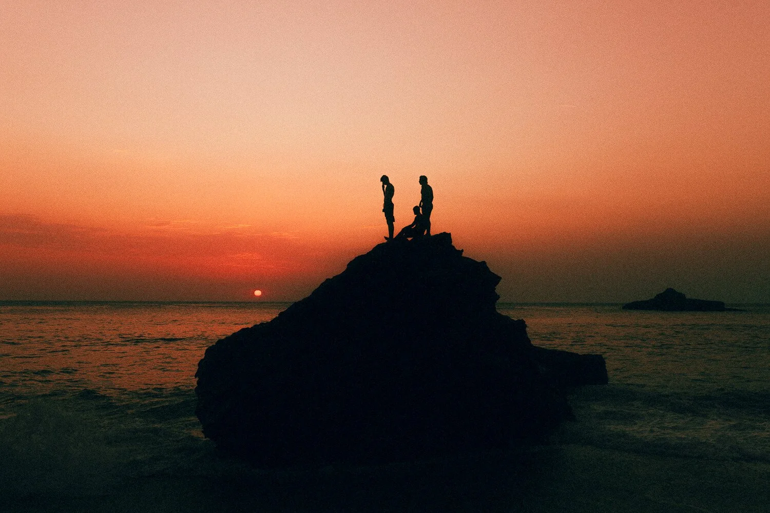 Silhouettes of three people standing on a large rock by the ocean during sunset, with the sun on the horizon and a pink-orange sky.