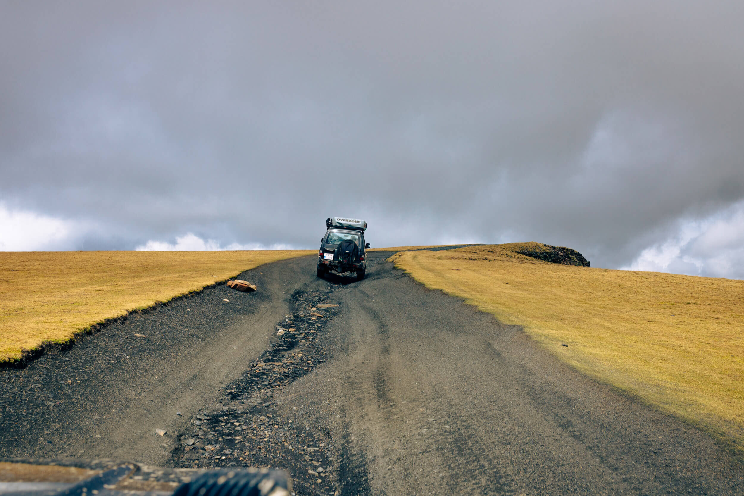 Une voiture sur une route de terre entourée de prairies sèches sous un ciel nuageux.