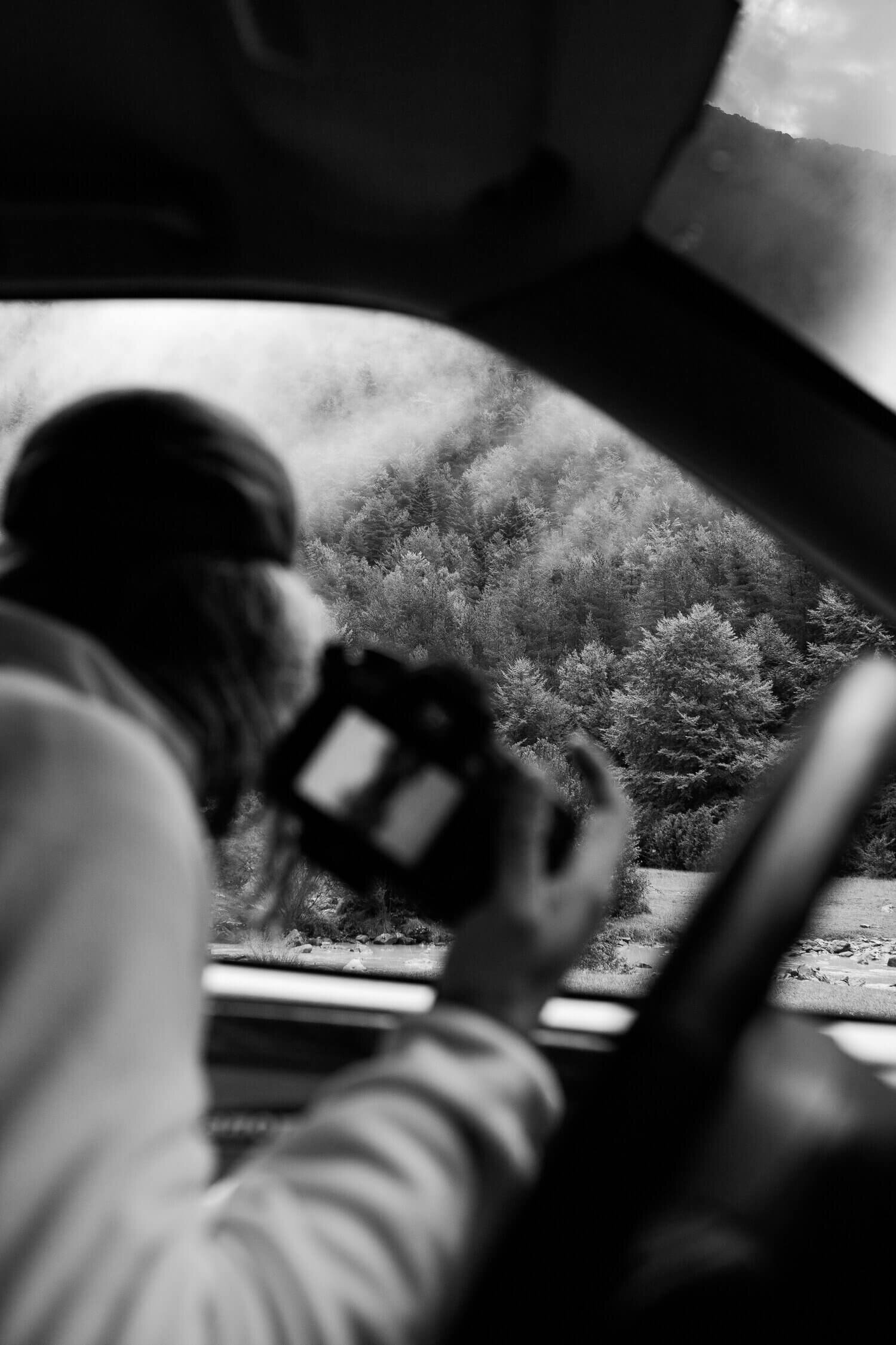 Personne dans une voiture regardant à travers la fenêtre avec un appareil photo en main, face à un paysage boisé montagneux et brumeux.