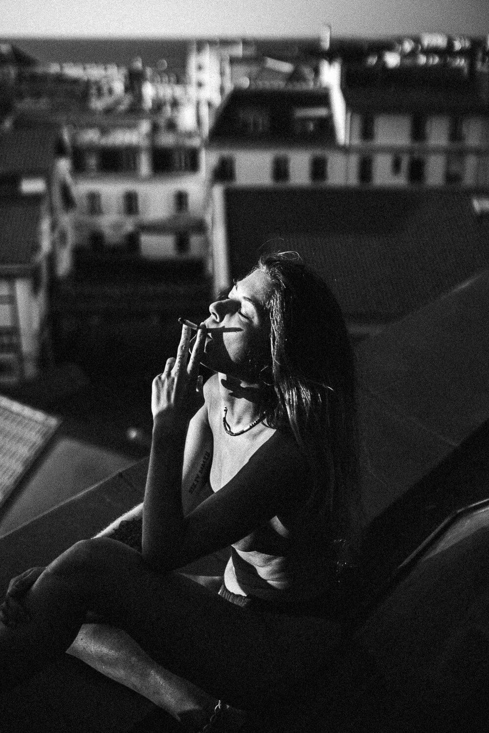 A woman with long dark hair and tattoos sitting on a rooftop at night, smoking a cigarette with city buildings in the background.
