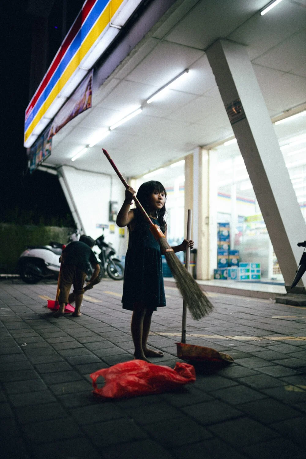Une jeune fille tenant un balai devant une station-service la nuit, avec un homme pliant des objets derrière elle et un scooter garé à côté.