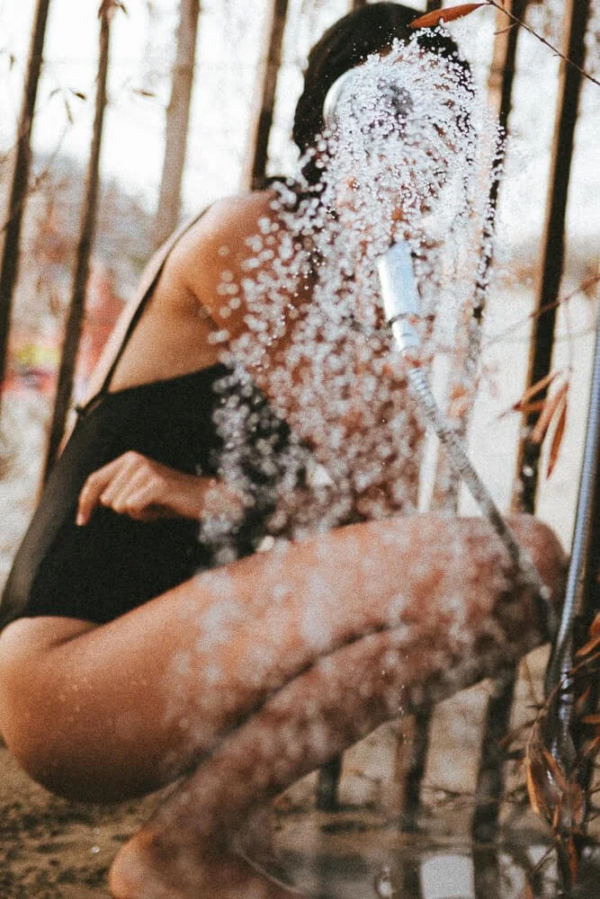 A woman with dark hair wearing headphones is taking a selfie through a glass with water droplets on it, outdoors among trees with brown leaves.