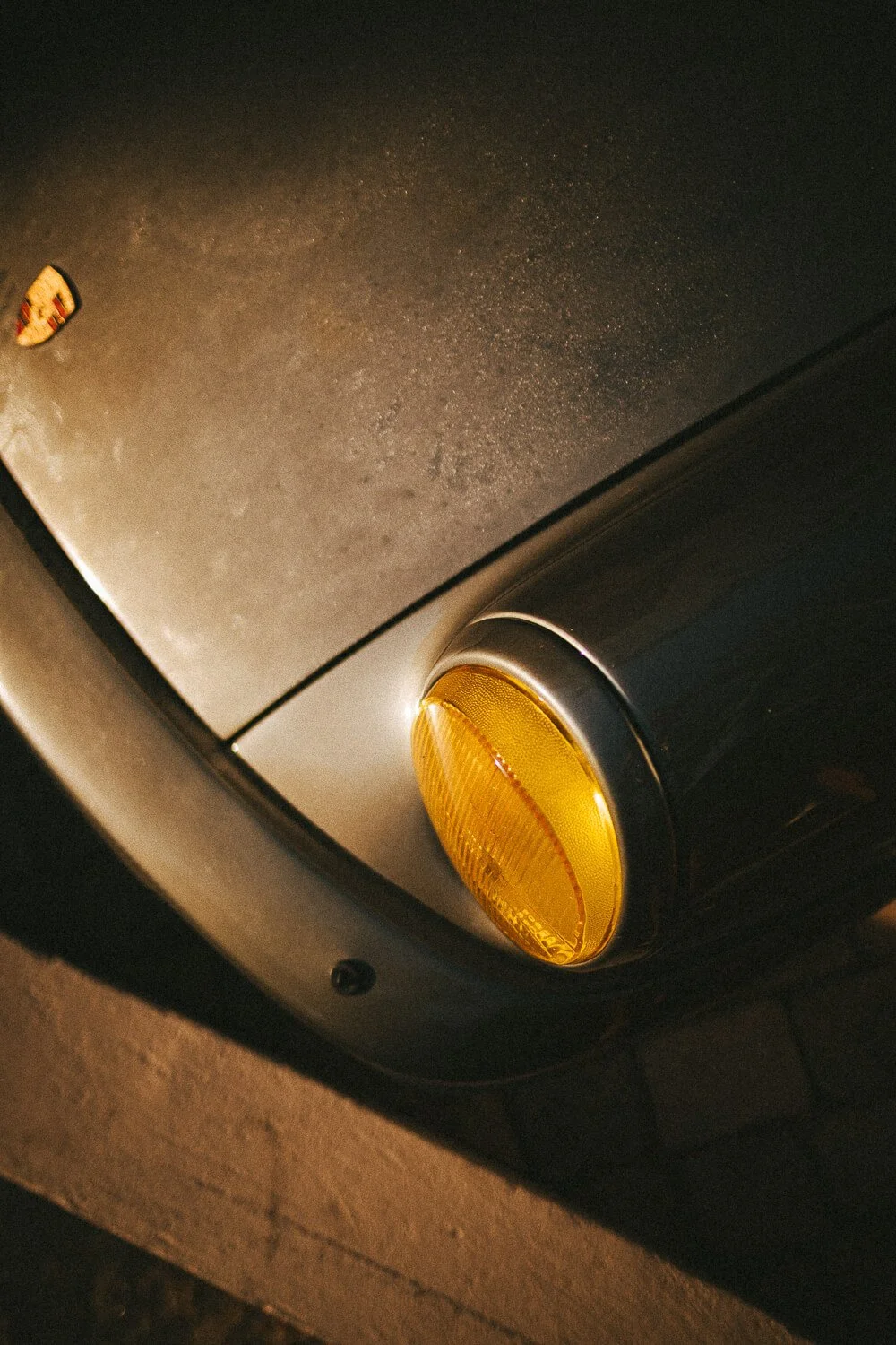 Close-up of the front corner of a vintage black Porsche car, showing the yellow turn signal light and part of the hood with a Porsche emblem visible.