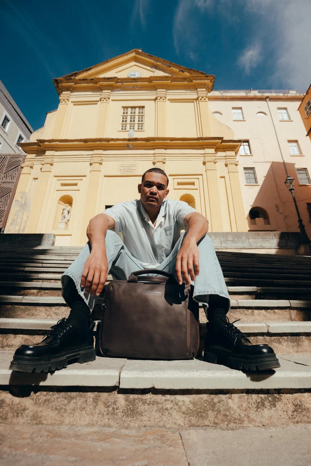 A young man sitting on stairs in front of a yellow church, wearing a white shirt and black boots, with a brown bag in front of him, under a clear blue sky.