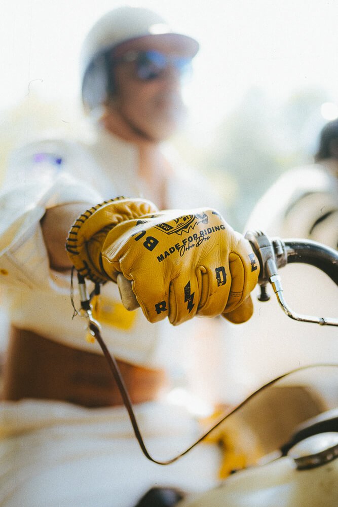 Close-up of a person wearing yellow racing gloves gripping the handlebar of a motorcycle during daytime. The person's face is blurred with sunglasses and a helmet.