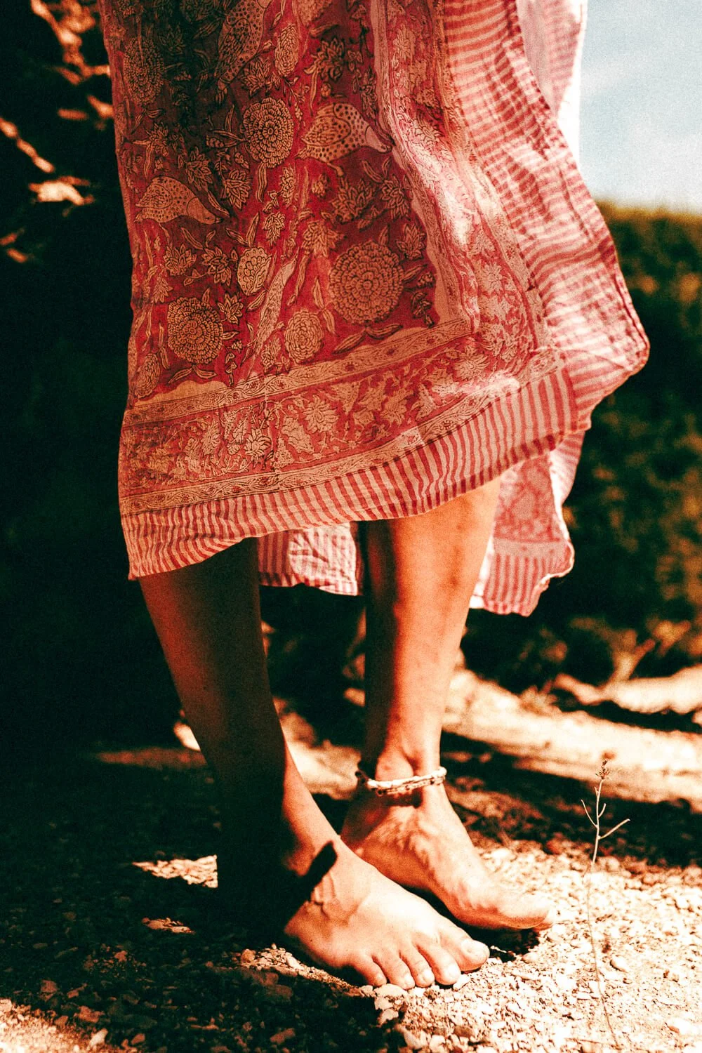A person standing barefoot on a dirt ground, wearing a pink and white patterned dress with floral and striped designs, along with an ankle bracelet. The photo is taken outdoors in natural light.