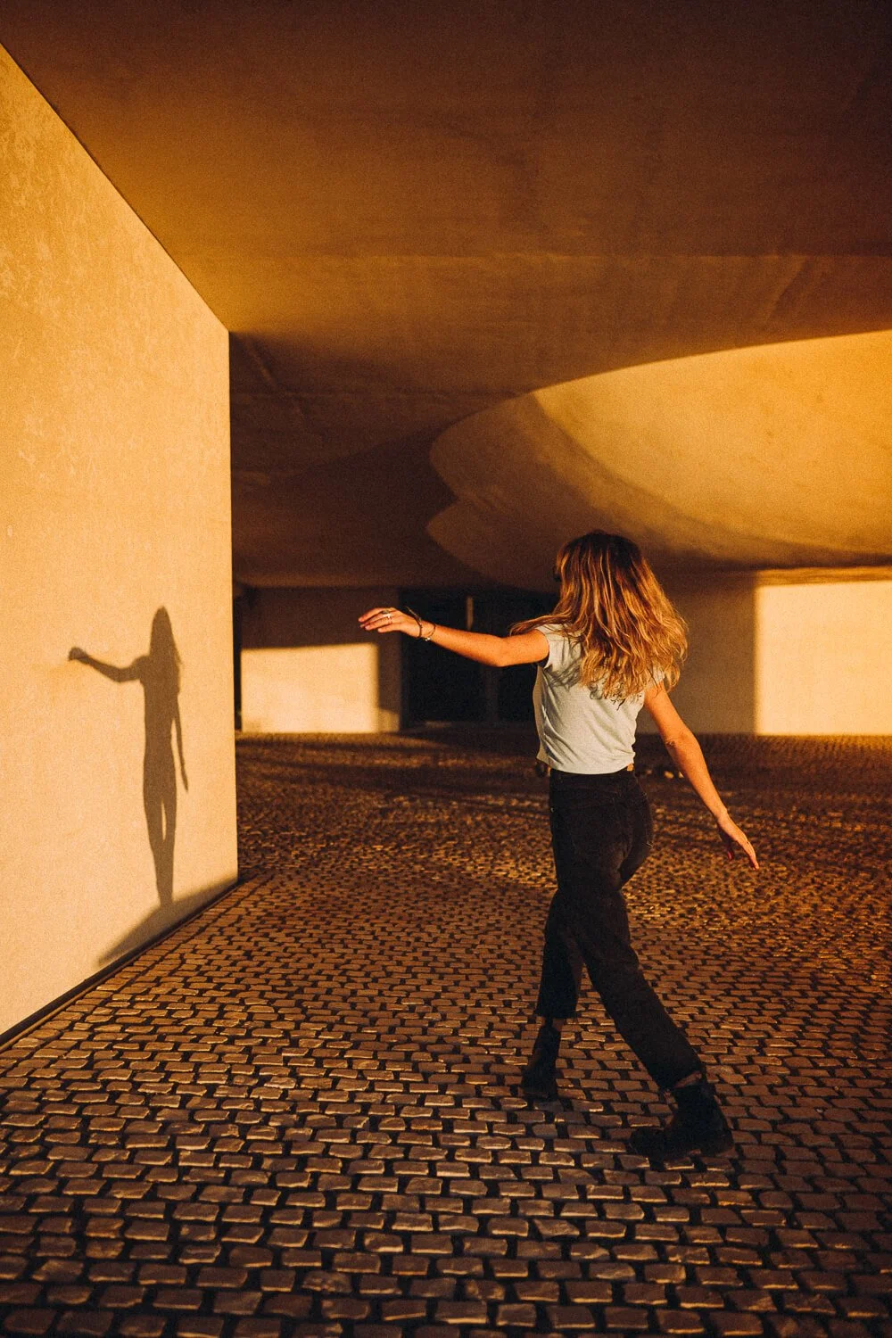 A woman dancing outdoors at sunset, casting a shadow on a beige wall, with a cobblestone ground and modern architectural structure overhead.