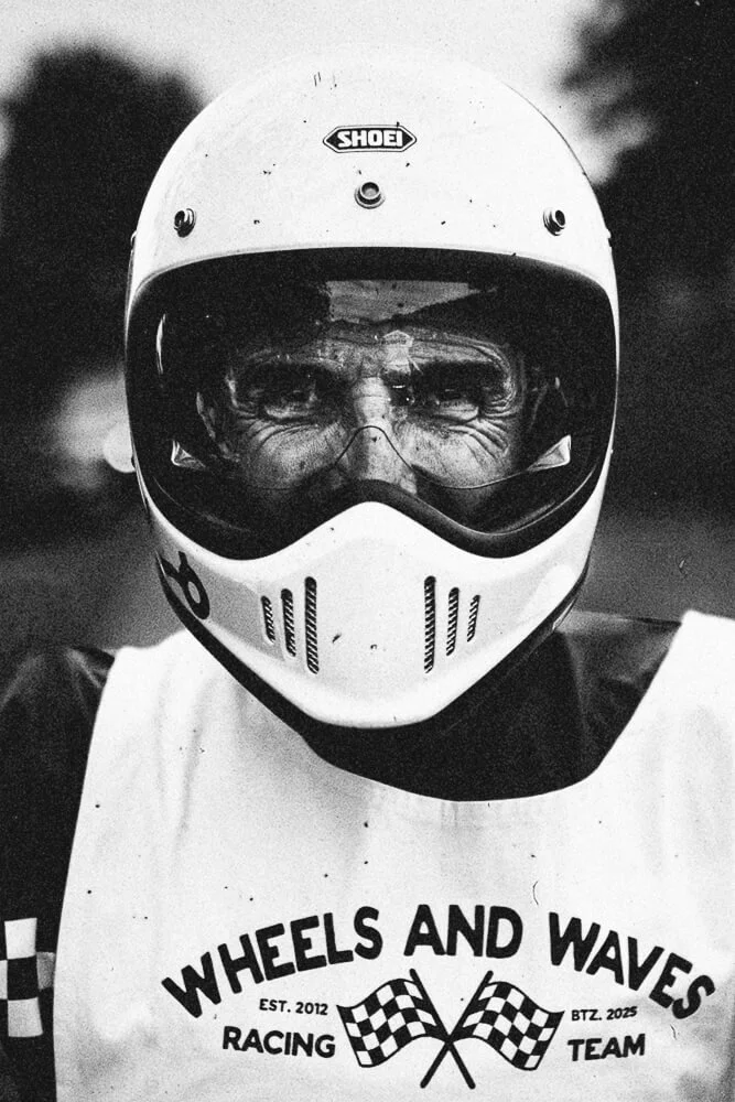 A person wearing a racing helmet and a vest with the logo "Wheels and Waves Racing Team" and checkered flags, standing outdoors.
