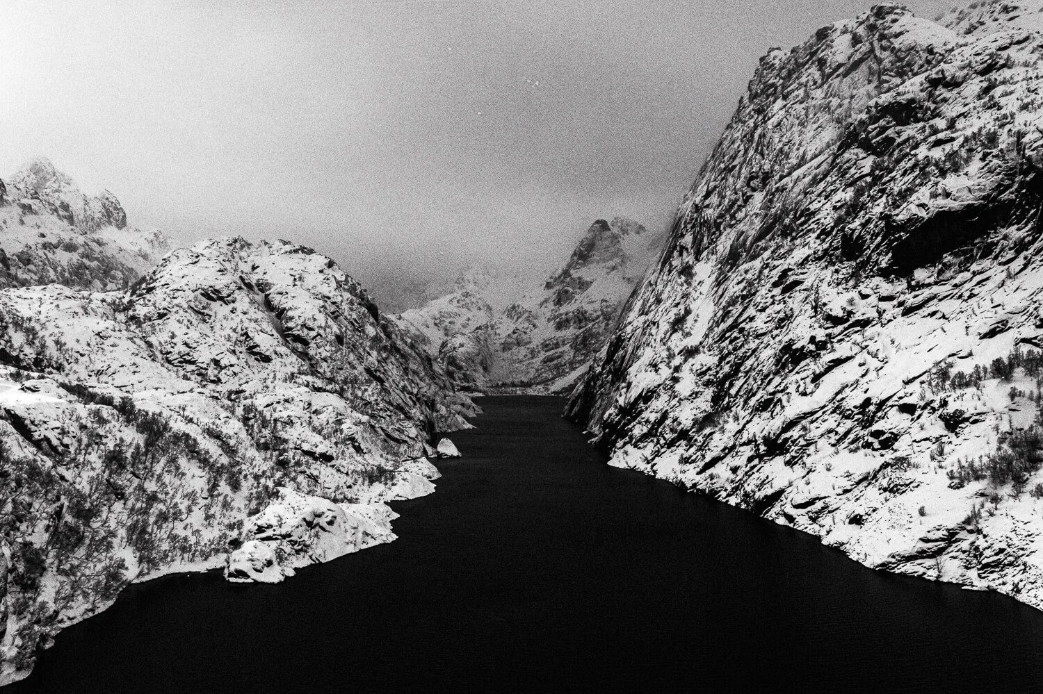 Vallée enneigée avec un lac noir au centre, entourée de montagnes rocheuses couvertes de neige, sous un ciel nuageux.