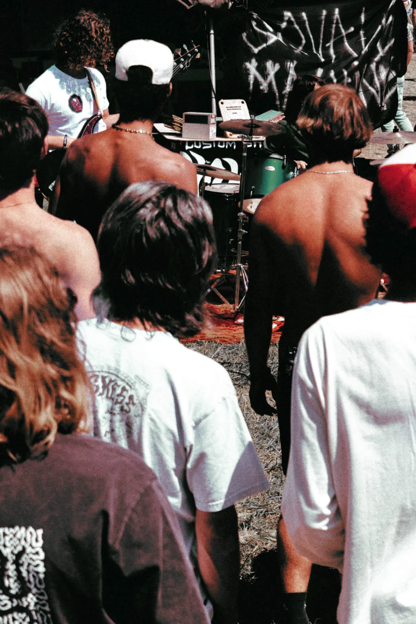 A live outdoor concert with people watching a band perform on stage at night, featuring musicians playing guitars and drums.