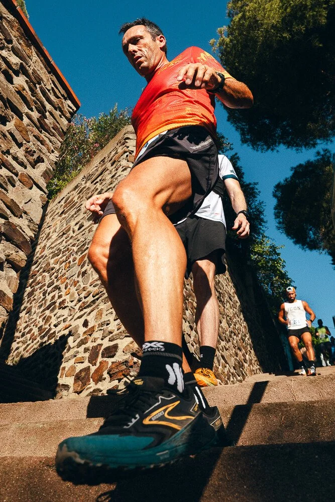 A man in a red shirt and black running shorts descending a set of outdoor stone stairs during a race or running event, with two other runners behind him and a stone wall on the side, under a clear blue sky with trees in the background.