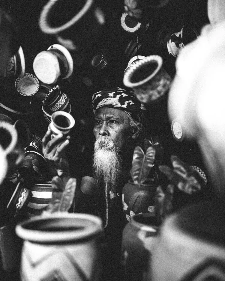 Un homme âgé avec une longue barbe et un bandana, entouré de pots en terre cuite, dans un environnement sombre.