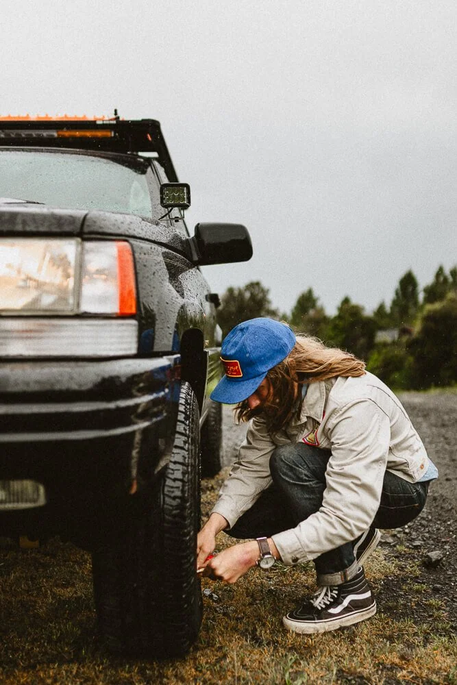 Person changing a flat tire on a black SUV in an outdoor setting on a rainy day.