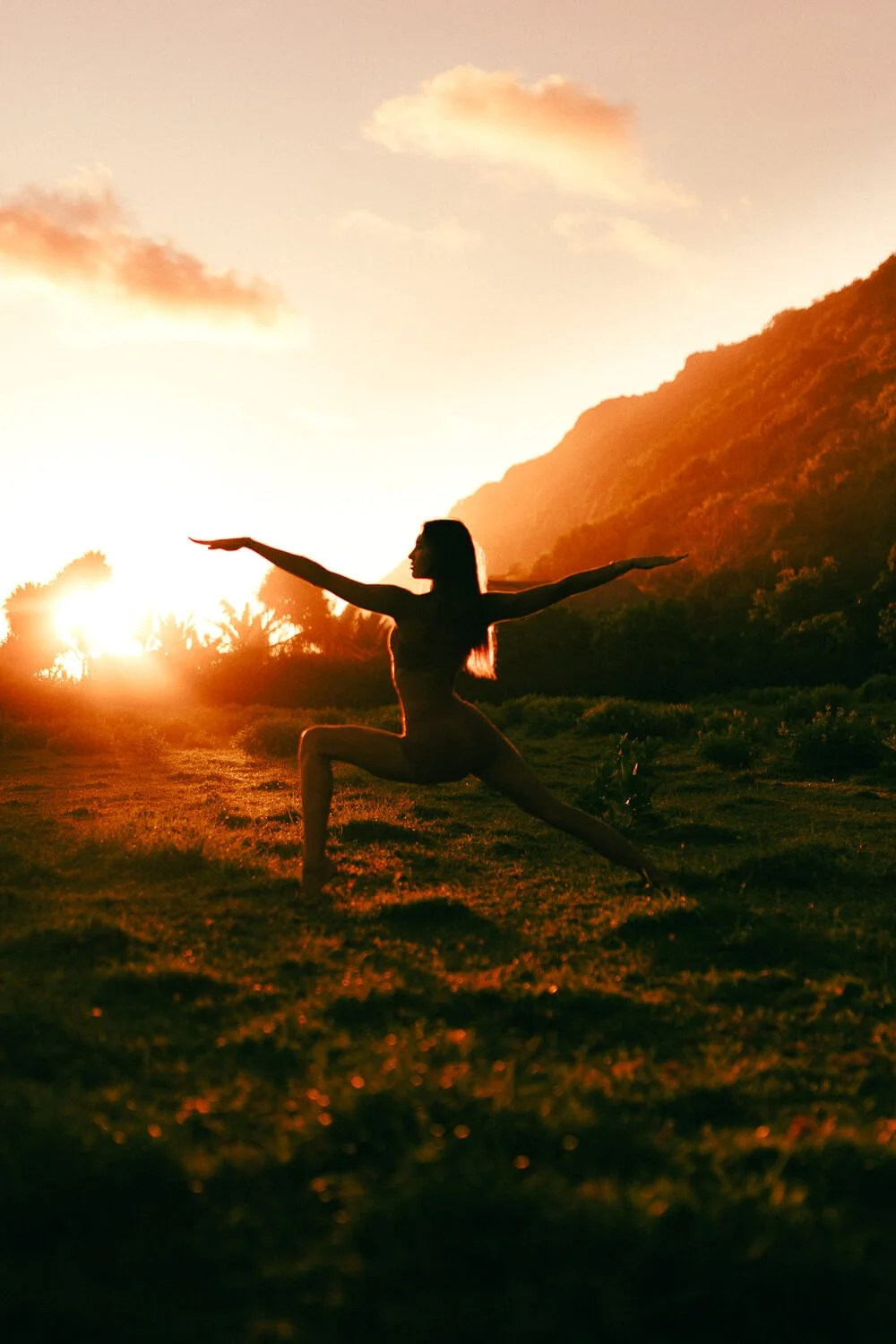 A woman practicing yoga outdoors during sunset, surrounded by mountains and lush greenery.