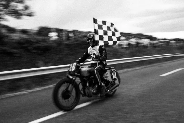 A motorcycle racer riding on a race track, holding a checkered flag, with motion blur in the background.