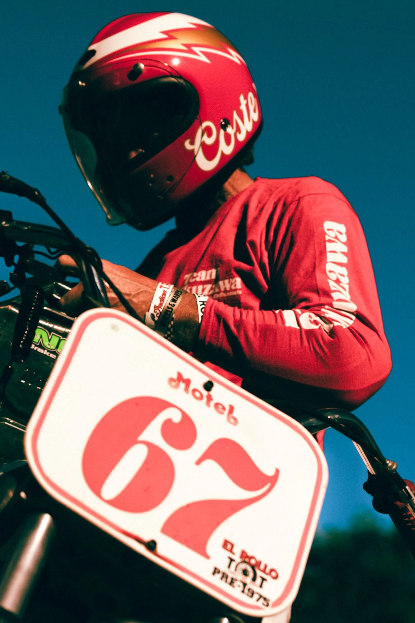 A person wearing a red helmet with the Coca-Cola logo, a red shirt with various logos, and wristbands, sitting on a motorcycle or bike with a blurred sign in the foreground and a clear blue sky in the background.